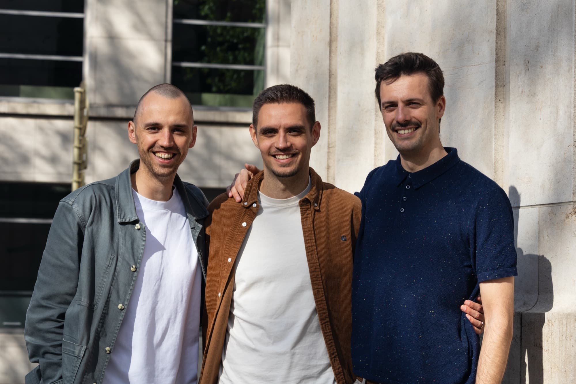 The three Lagotta founders, Max Rymen, Justin Bériot and Jeroen Thierens, smiling together in front of a stone facade in the sun